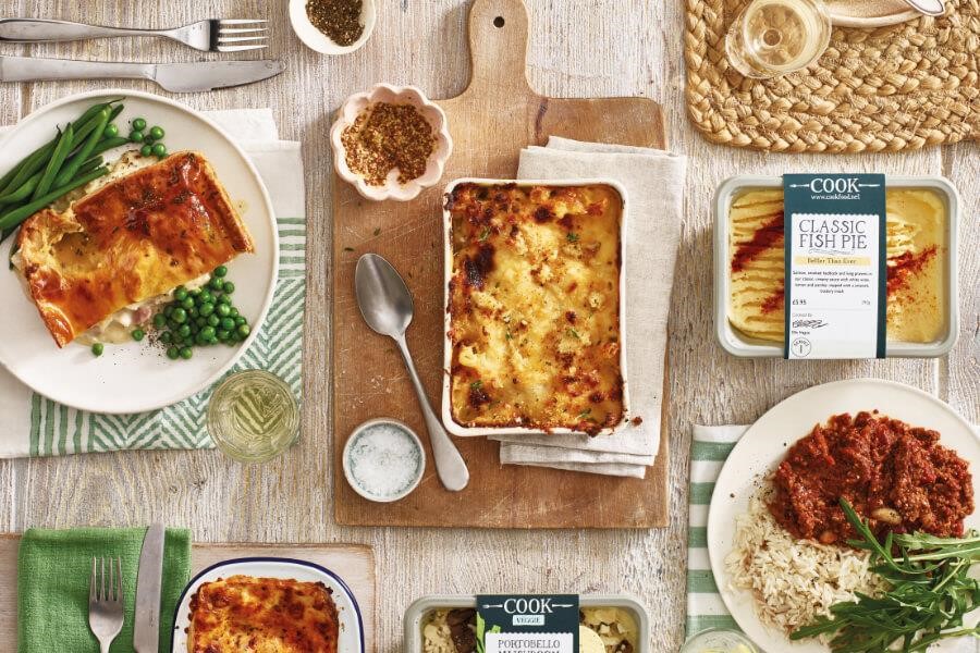 An assortment of COOK meals laid out on a wooden table, including a steak pie with peas, a creamy bake, and a tomato pasta dish.