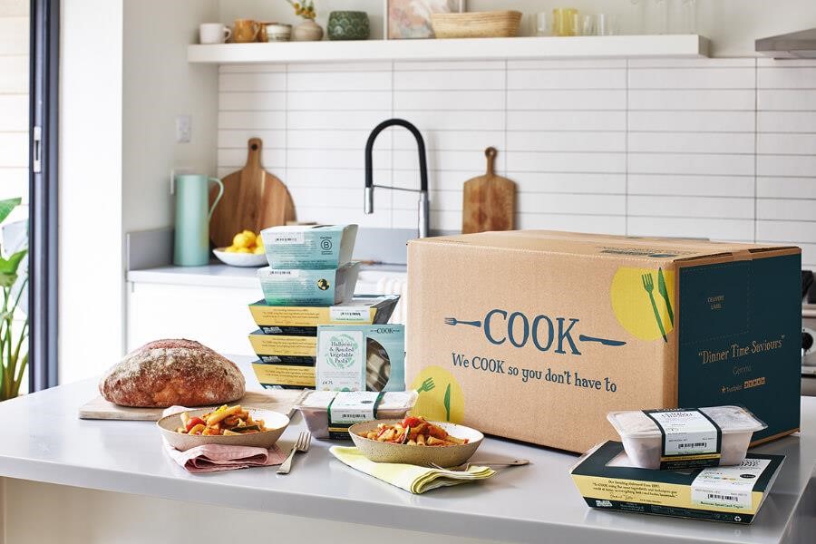 A COOK delivery box on a kitchen countertop surrounded by frozen meals, a loaf of bread and bowls of pasta.